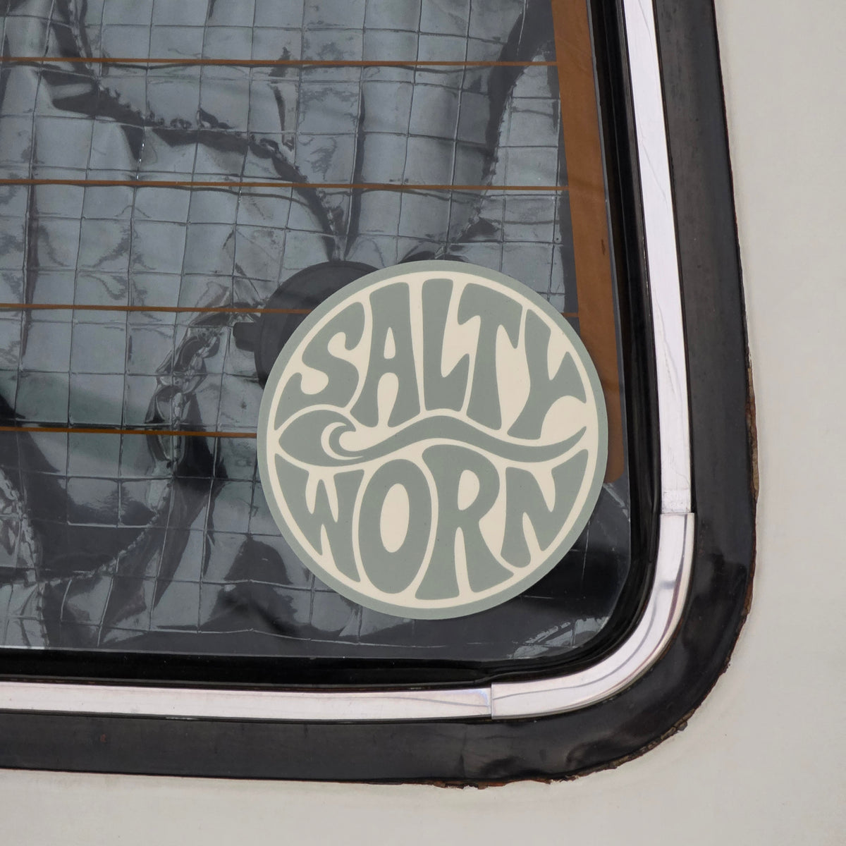 Salty Worn round retro logo sticker in sage green and cream displayed on a car rear window.